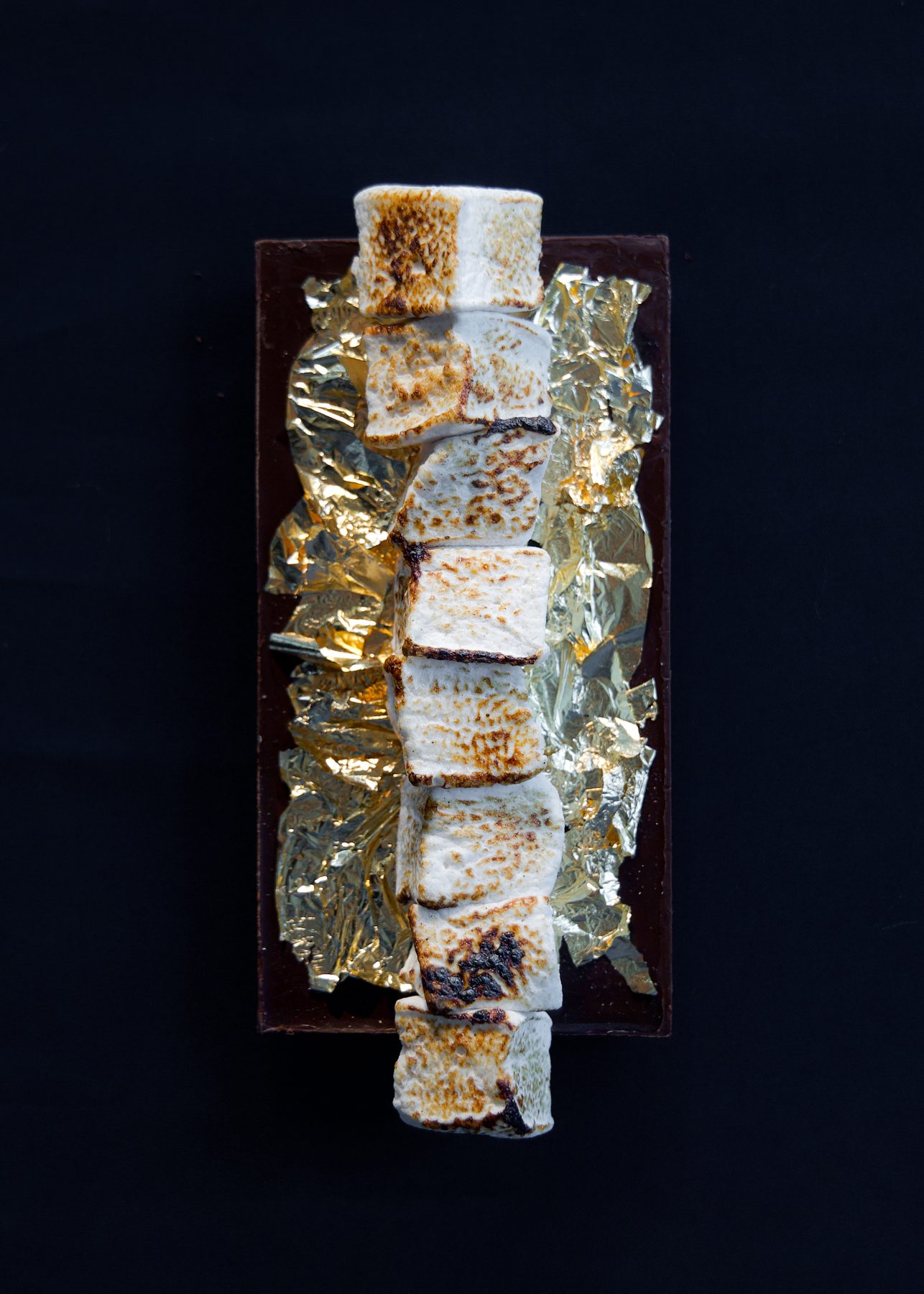 Toasted marshmallows stacked on dark chocolate and gold leaf against a dark backdrop