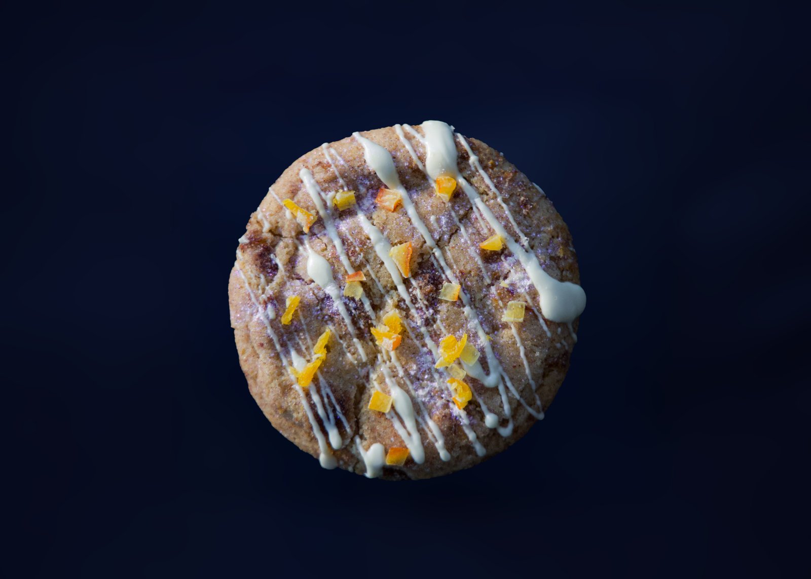 A sugared cookie with white chocolate drizzle and candied orange peel against a dark blue backdrop