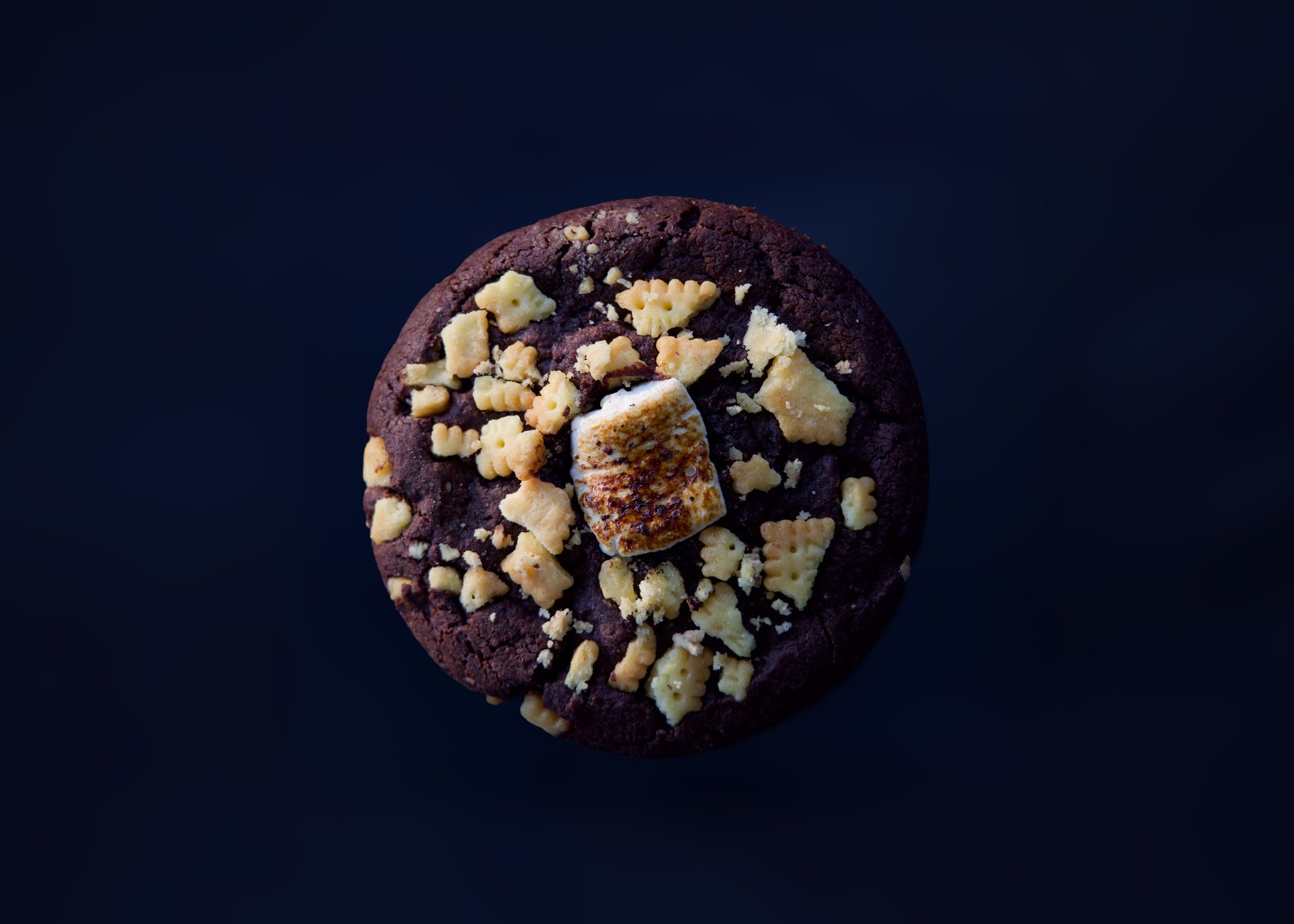 A dark chocolate cookie topped with biscuit pieces and a toasted marshmallow against a dark blue backdrop